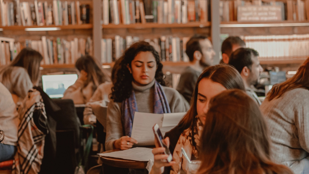 A university library. There are tables with lots of students sitting around with large bookshelves in the background.